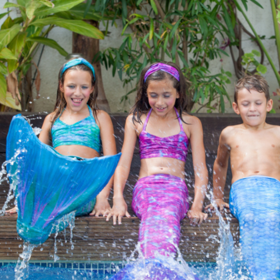 Three children looking really happy splashing in the pool with their mermaid fins.